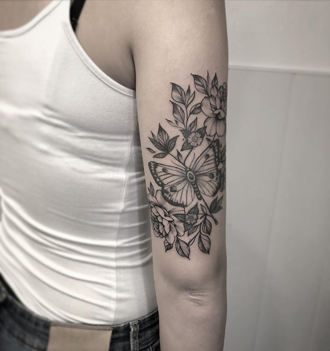large black and grey tattoo on woman's upper arm of butterfly with flowers and leaves around it