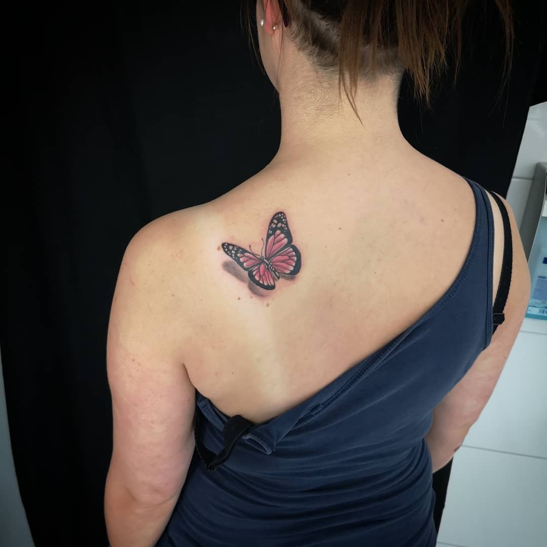 medium-sized color tattoo on woman's shoulder of pink flying butterfly with shadow