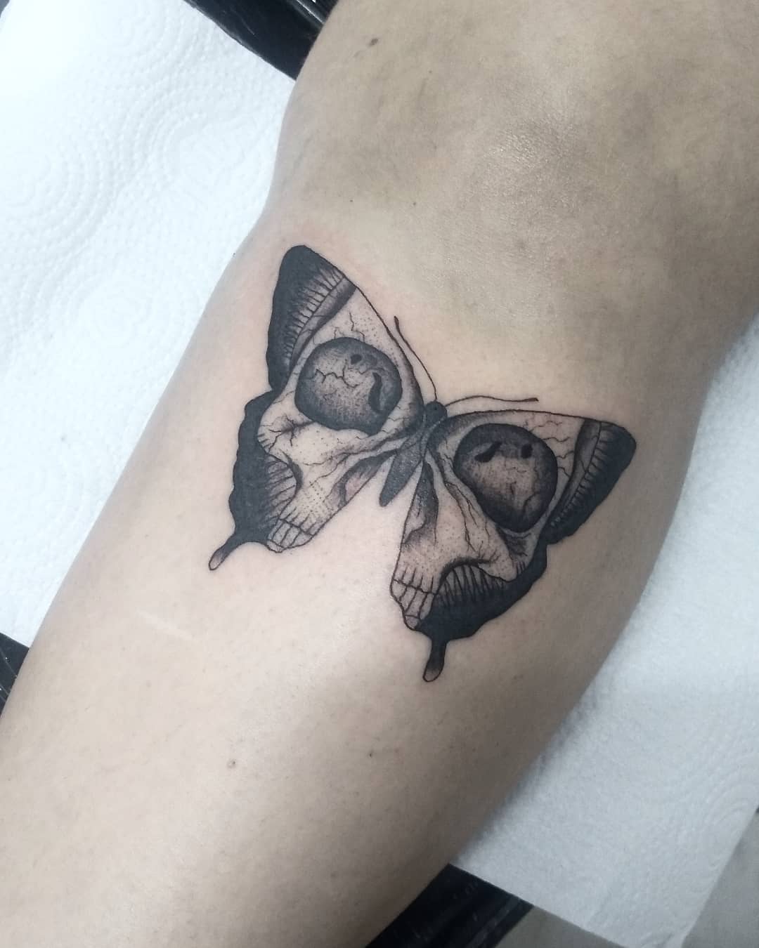 medium-sized black and grey tattoo on man's lower leg of a surrealistic butterfly with skull face in its wings