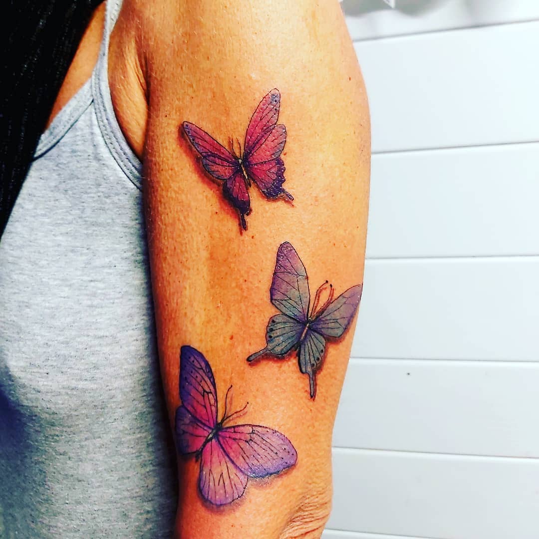large color tattoos on woman's upper arm of three realistic pink and blue butterflies