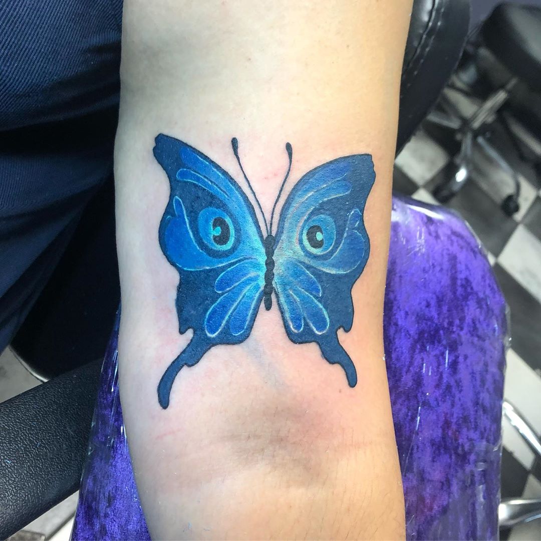 large color tattoo on woman's upper arm of blue butterfly with eyes on its wings